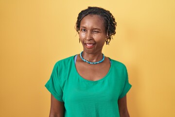 African woman with dreadlocks standing over yellow background winking looking at the camera with...