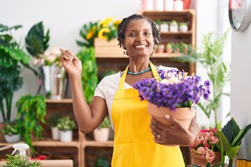 African woman with dreadlocks working at florist shop holding plant smiling happy pointing with...