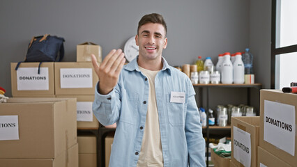Smiling young hispanic man confidently gestures 'come' while volunteering at a charity center,...