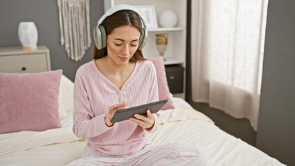 A woman in pink pajamas uses a tablet in a cozy bedroom while wearing wireless headphones.
