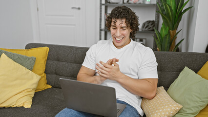 A cheerful young hispanic man with curly hair sitting on a gray couch using a laptop in a cozy living room.