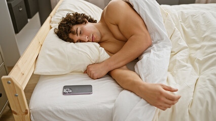 Obraz na płótnie Canvas A young hispanic man with curly hair sleeps shirtless in a cozy bedroom, with a smartphone lying beside him.