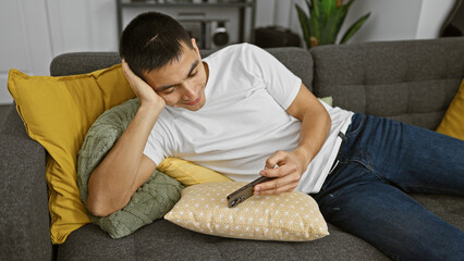 A relaxed young hispanic man leisurely uses a smartphone while lounging on a cozy couch with decorative pillows in a modern living room.