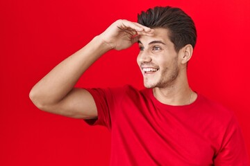 Fototapeta na wymiar Young hispanic man standing over red background very happy and smiling looking far away with hand over head. searching concept.