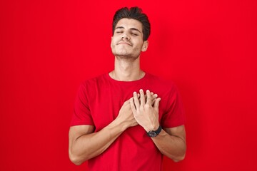 Young hispanic man standing over red background smiling with hands on chest with closed eyes and grateful gesture on face. health concept.