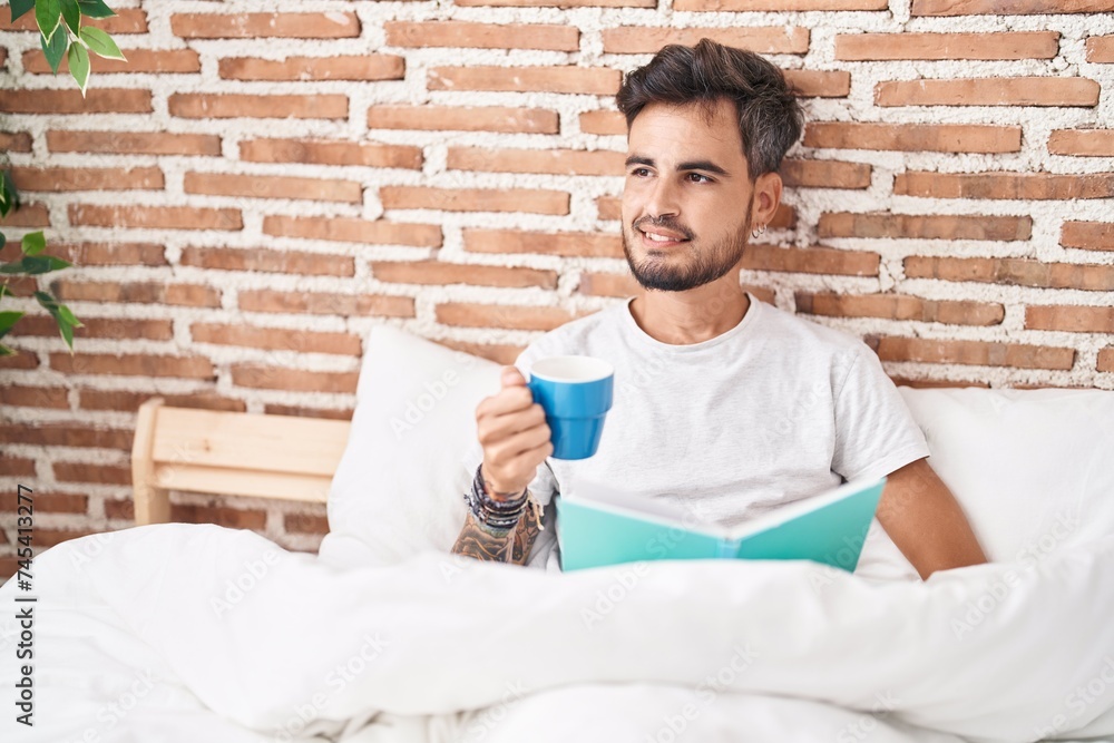 Wall mural Young hispanic man drinking cup of coffee reading book at bedroom