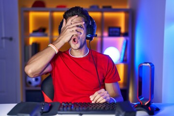Young hispanic man playing video games peeking in shock covering face and eyes with hand, looking through fingers with embarrassed expression.