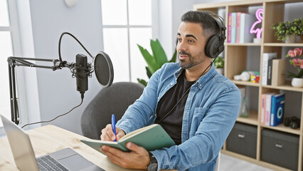 A bearded young hispanic man podcasts indoors with headphones, a microphone, and a laptop, looking thoughtful.
