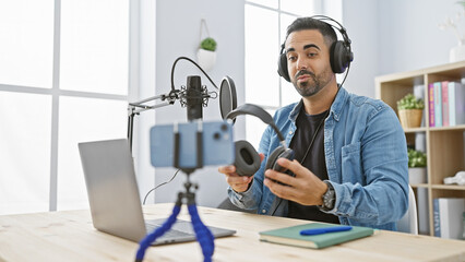 Hispanic man with beard hosting podcast in modern studio, laptop and microphone on table.