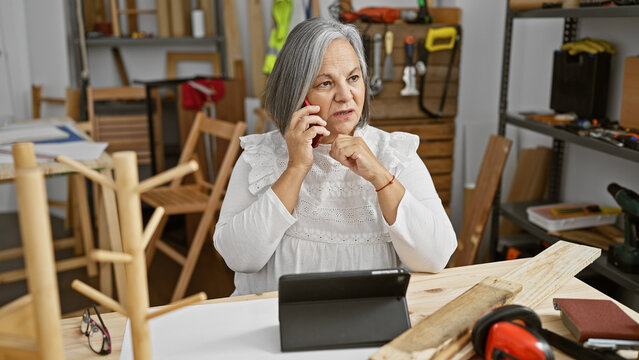 Mature woman conversing on phone in workshop surrounded by woodworking tools.