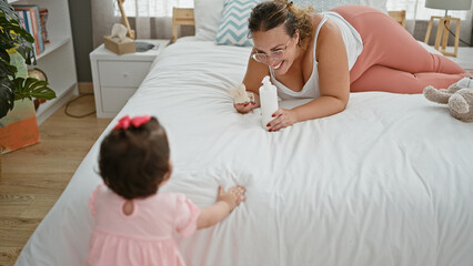 Obraz na płótnie Canvas Confident mother and cheerful daughter enjoying playtime with toys, sitting on a comfy bed in a sunlit bedroom, expressing joy and love with warm smiles