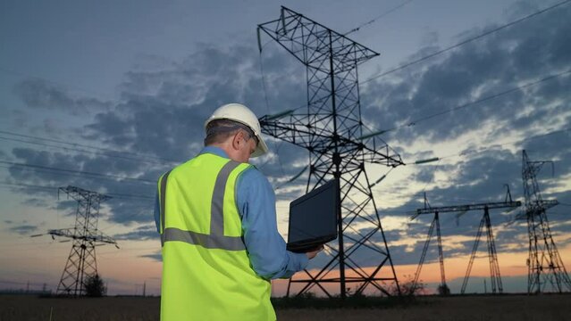 Technician holds laptop walking to modern power transmission lines in dusk rural field backside view. Man engineer in helmet works on laptop at power generation substation in evening country meadow