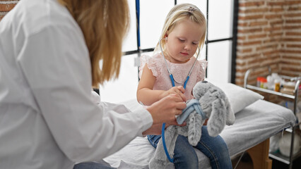 Fototapeta premium Cute little girl patient, teddy bear in arm, gets a playful heart checkup with stethoscope by caring pediatrician doctor at indoor clinic - childhood healthcare and medical care made fun