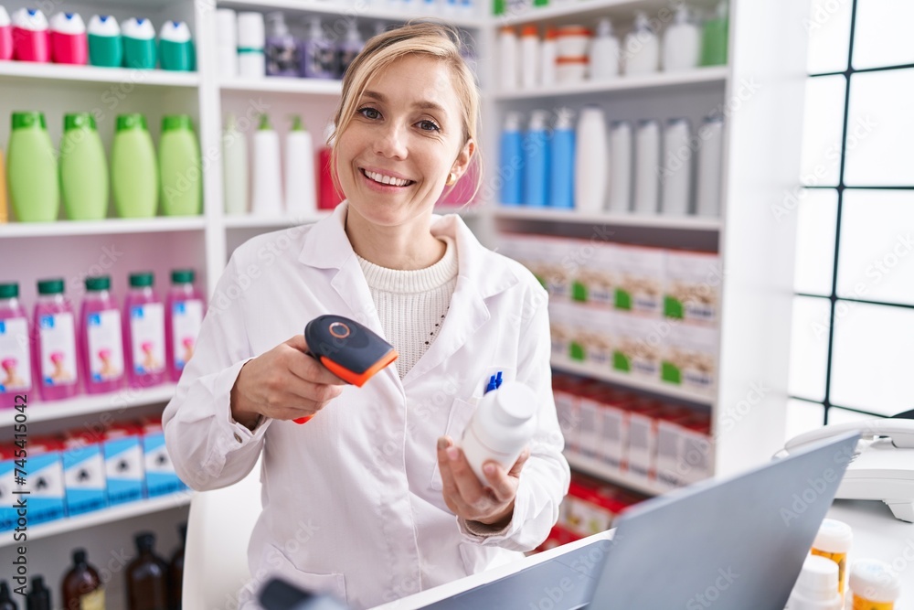 Wall mural young blonde woman pharmacist scanning pills using laptop at pharmacy