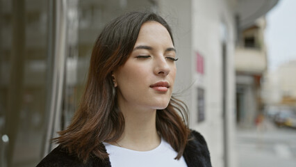 A calm hispanic woman with a subtle smile stands outdoors on a city street, exuding confidence and beauty.