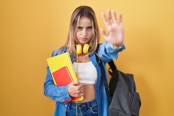 Young blonde woman wearing student backpack and holding books doing stop sing with palm of the hand. warning expression with negative and serious gesture on the face.