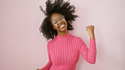 A joyful african woman in glasses and a pink sweater celebrating indoors with a fist pump gesture