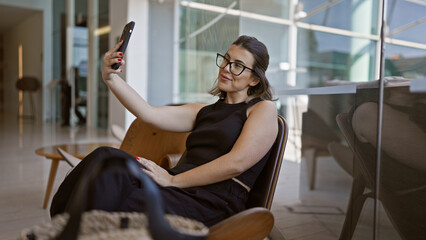 Confident hispanic woman with glasses having fun, taking a radiant selfie while casually sitting in a modern hotel hall