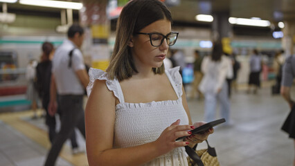 Stunning hispanic woman, glasses on, standing at underground railroad station, anticipating her subway journey, engrossed in her phone