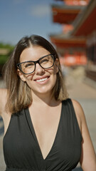 Confident hispanic woman, beautiful and joyful, cheerfully posing with glasses on, standing and smiling on kyoto's traditional streets, radiating natural and carefree latin expression.