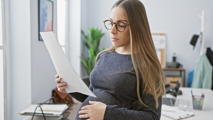 A pregnant hispanic woman reads a document in an office, portraying maternity in the workplace.