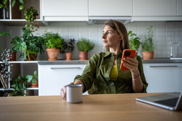 Pensive woman freelancer working on laptop at home, having tea, coffee break, sitting at table with smartphone, looking at window, dreaming, thinking of news read. Mental relaxation after hard job