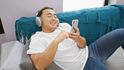 Smiling, confident young chinese man happily engrossed in watching an online video on his sleek smartphone, relaxing indoor on a comfy sofa in his apartment's intimate living room