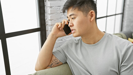 A handsome asian man is thoughtfully looking away indoors, seated in a modern apartment living room, portraying casual, contemporary lifestyle.