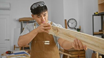 A focused young asian man examines a wooden plank at his well-equipped indoor carpentry workshop.