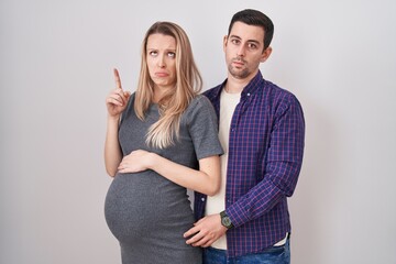 Young couple expecting a baby standing over white background pointing up looking sad and upset,...