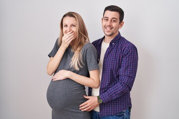 Young couple expecting a baby standing over white background laughing and embarrassed giggle...