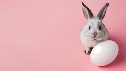 Petit lapin posant à côté d'un œuf blanc sur un fond rose pastel - avec espace pour texte - obrazy, fototapety, plakaty