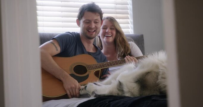 Happy couple, home and playing guitar with dog for love and bonding in bed with pet. People with animal, playful husky and fun with musical instrument, time together for trust, commitment and art