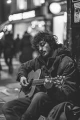 A man, VetalVit, an indie folk singer-songwriter, sitting on a bench while strumming an acoustic guitar - obrazy, fototapety, plakaty
