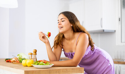 Portrait of positive girl in purple nightgown tasting vegetable salad at kitchen