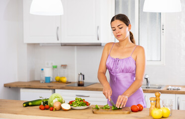 Obraz na płótnie Canvas Young woman wearing nightie using kitchen knife to slice fresh vegetables for salad. Preparing food at home.
