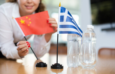 Young female assistant setting up greek and chinese flags for international negotiations