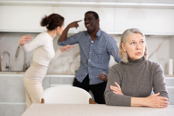Upset old-aged woman sitting in the kitchen and middle-aged man and woman arguing angrily standing...
