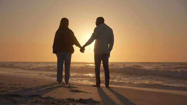 Rear View Of Casually Dressed Loving Young Couple Standing Beach Shoreline Holding Hands At Sunrise
