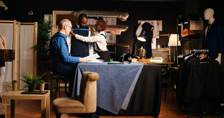 Customer working with senior tailor to design tailored suit in atelier, male client talking to...