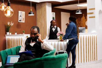 Young African American man hotel manager sitting in lobby talking by phone and taking notes,...