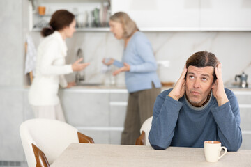 Old woman is quarreling with woman while sad man is sitting with them in kitchen