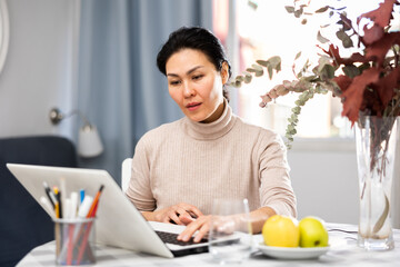 Fototapeta premium Asian woman sitting at table in apartment and using her laptop.