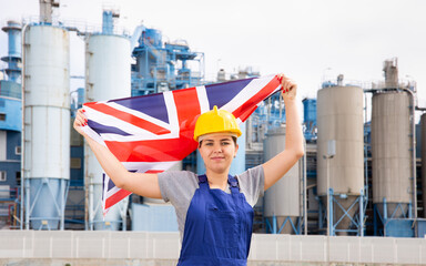 Young female engineer in helmet holding state flag of Great Britain while standing in front of big tanks at chemical plant 