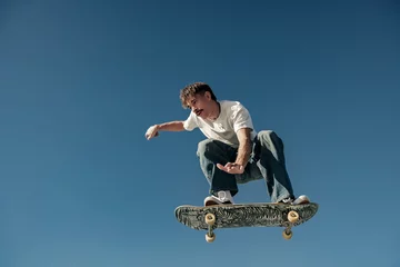 Gordijnen Active man doing tricks in the air on his skateboard at the skate park on blue sky background © Kostiantyn