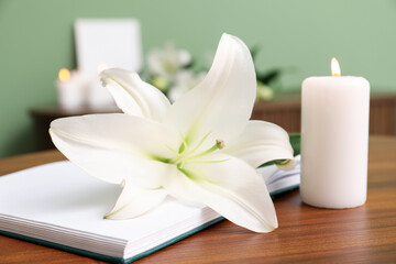 Beautiful lily flower, burning candle and book on wooden table in room, closeup