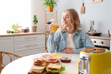 Young beautiful happy woman drinking orange juice with tasty sandwiches in kitchen