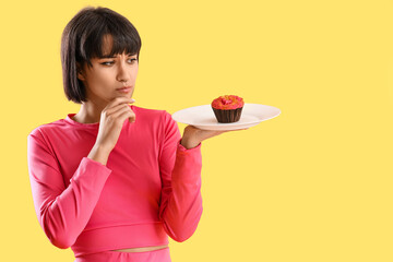 Thoughtful sporty young woman with muffin on yellow background. Diet concept