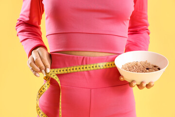 Sporty young woman with oatmeal measuring her waist on yellow background, closeup. Diet concept