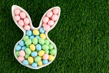 Pastel candy coated chocolate eggs in an Easter Bunny shaped white ceramic dish on a green grass...